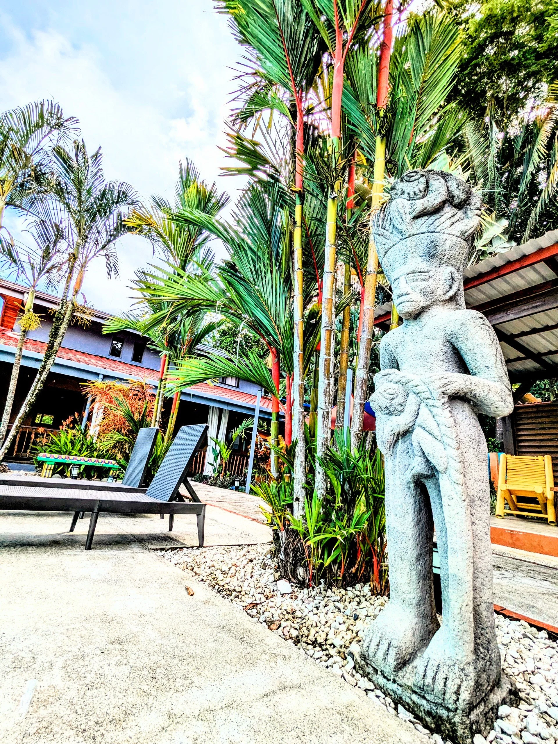 A stone sculpture of a person with a headpiece, standing outdoors among tropical plants, with a building and lounge chairs in the background.