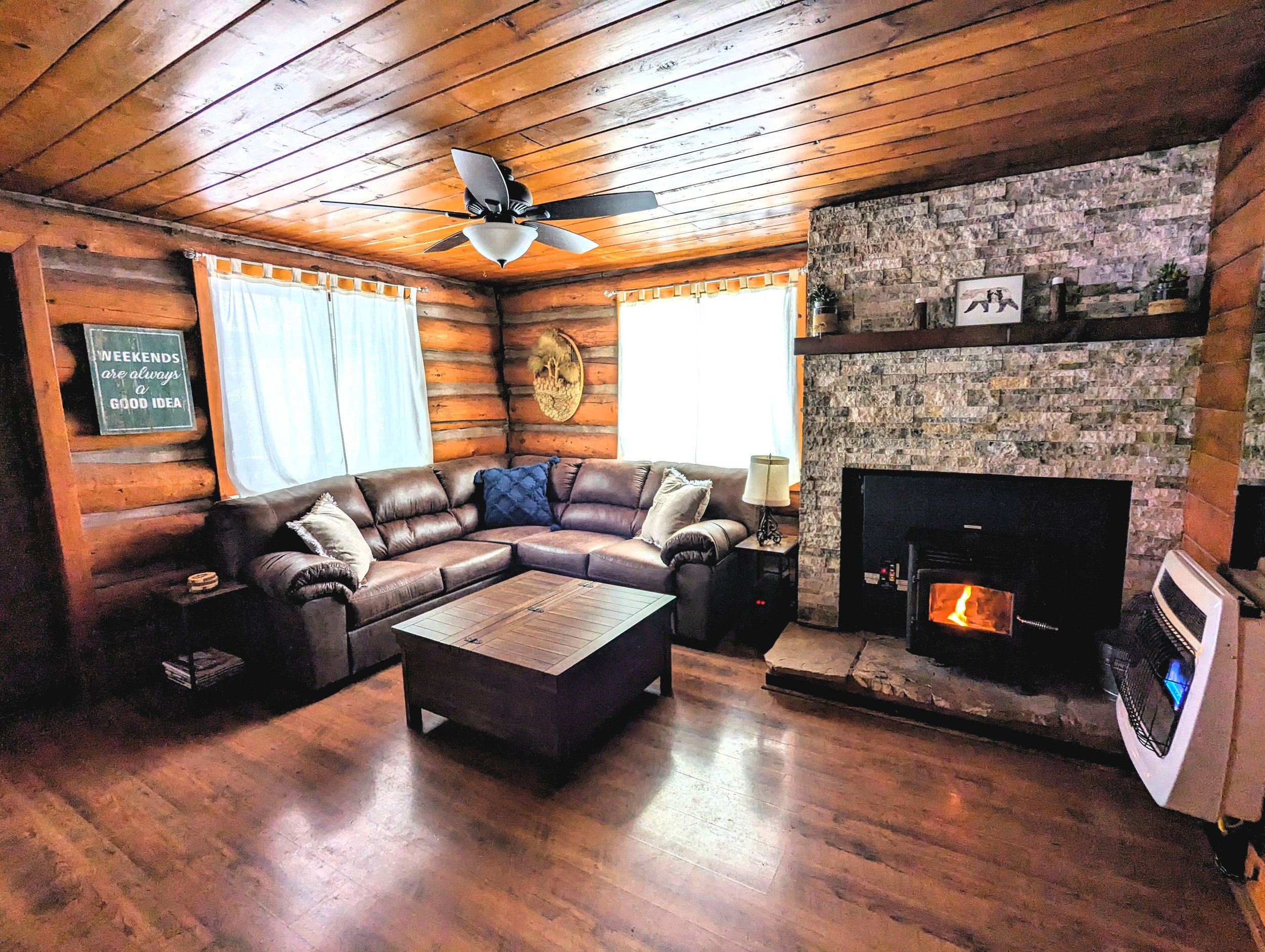 Cozy living room with wooden walls and ceiling, a leather sectional sofa with pillows, a wooden coffee table, a stone fireplace with a fire, a ceiling fan, and a heater.