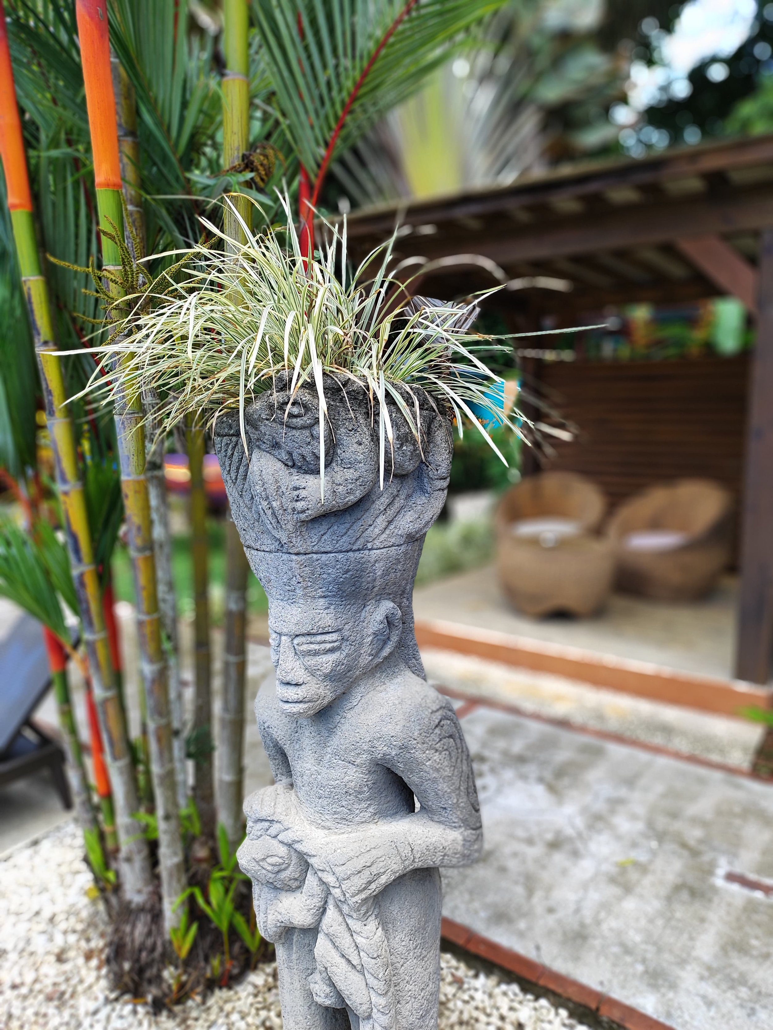 A concrete garden sculpture of a human figure with a flower pot on its head, containing plants, outdoors with bamboo and wooden patio furniture in the background.