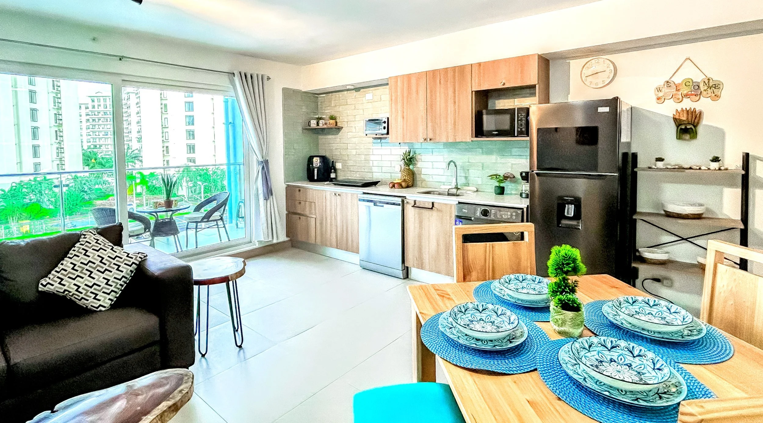 Open-concept living and kitchen area with a sliding glass door leading to a balcony with outdoor chairs. The living area features a black sofa with a black-and-white patterned pillow. The dining table is set with four plates and blue placemats, with a small green plant centerpiece. The kitchen has wooden cabinets, a white brick backsplash, stainless steel appliances, and decorative items on shelves and countertops.