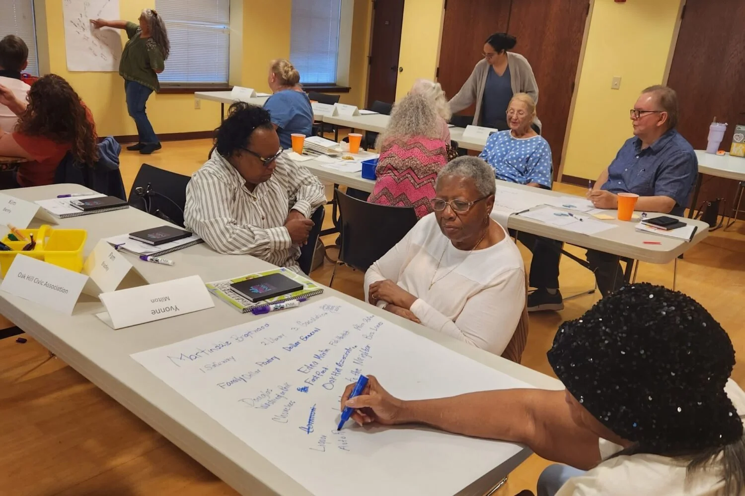 A group of diverse adults participating in a community meeting or workshop, some seated at tables with notebooks and name tags, others standing or writing on a large sheet of paper, in a room with yellow walls and wooden floors.