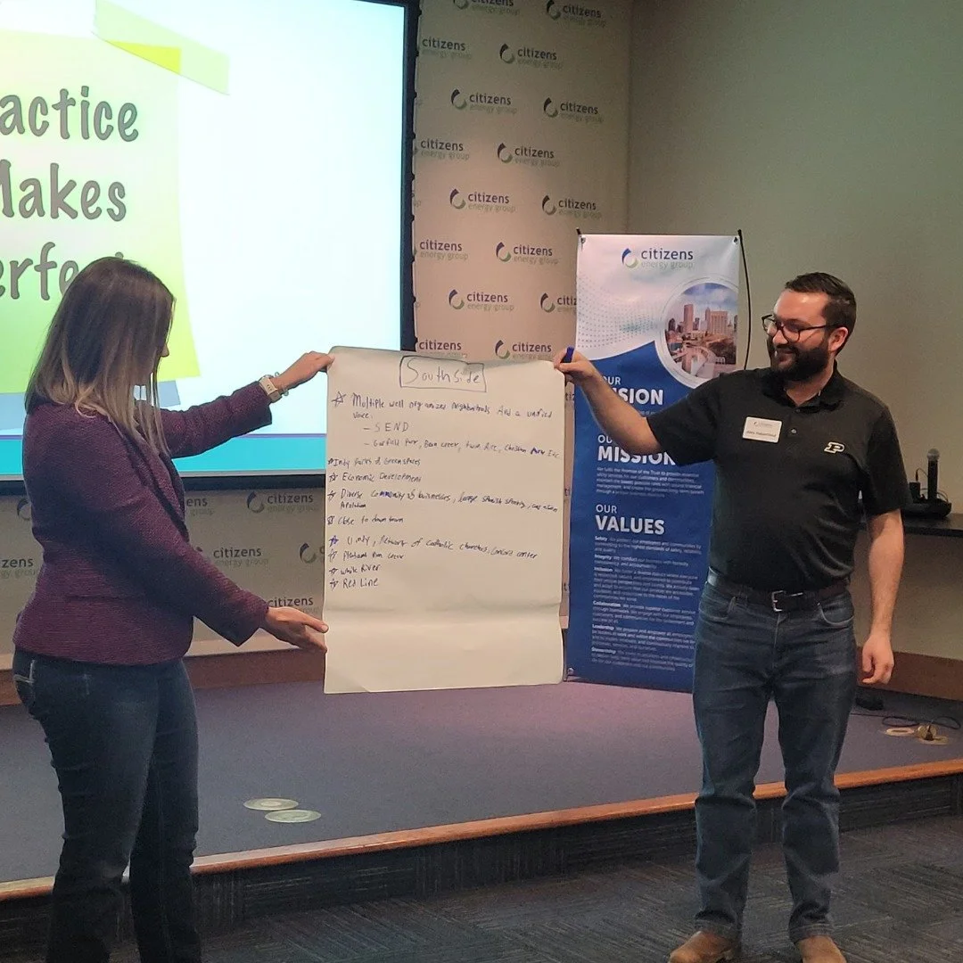 Two people standing in front of a presentation screen and banner, holding a large sheet of paper with handwritten notes, in a conference room.