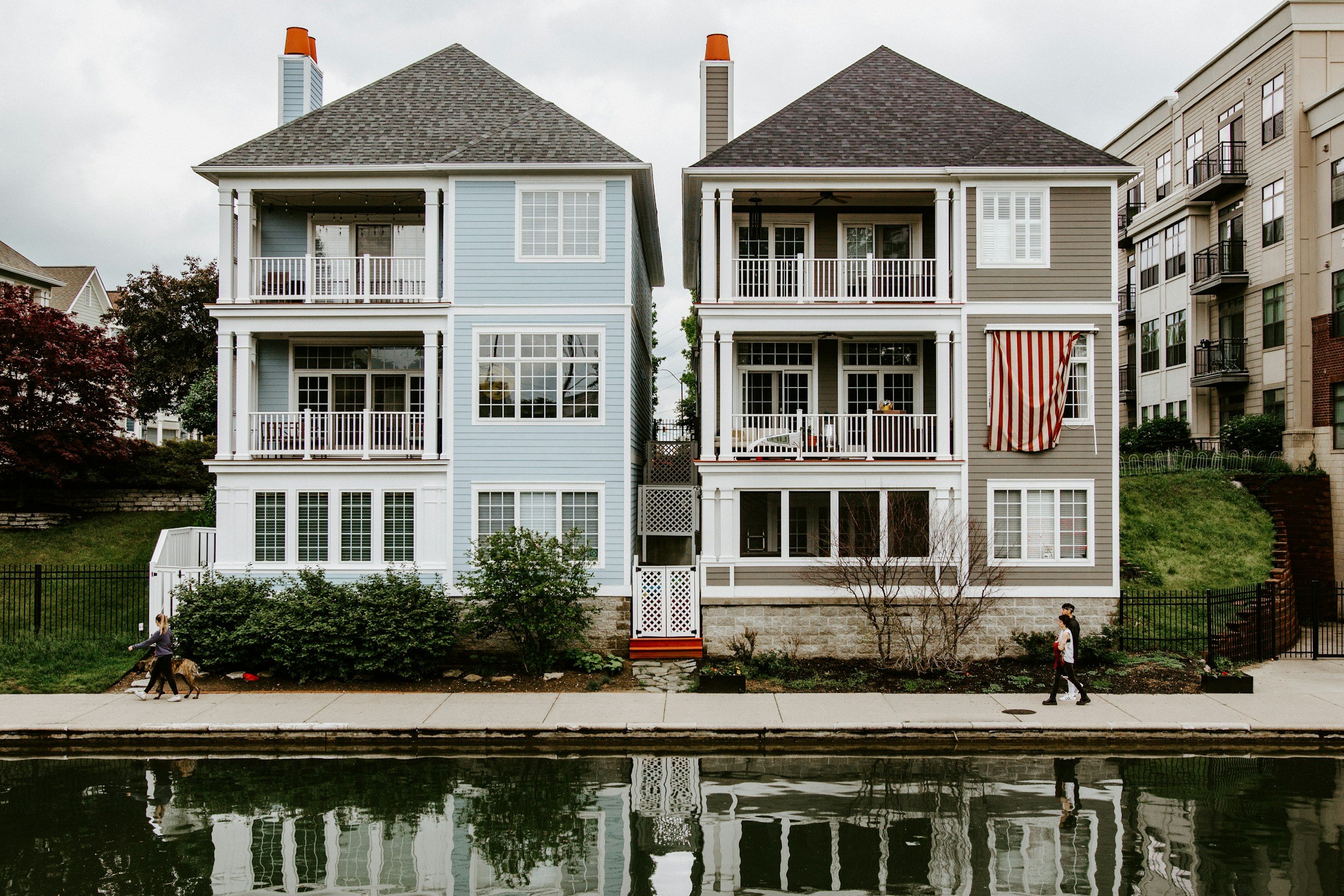 Two multi-story residential buildings with large windows and balconies, situated along a canal with a sidewalk in front. Two people are walking on the sidewalk, one with a dog. The buildings have different colors, one light blue and the other grey, with a fence and shrubbery in front.