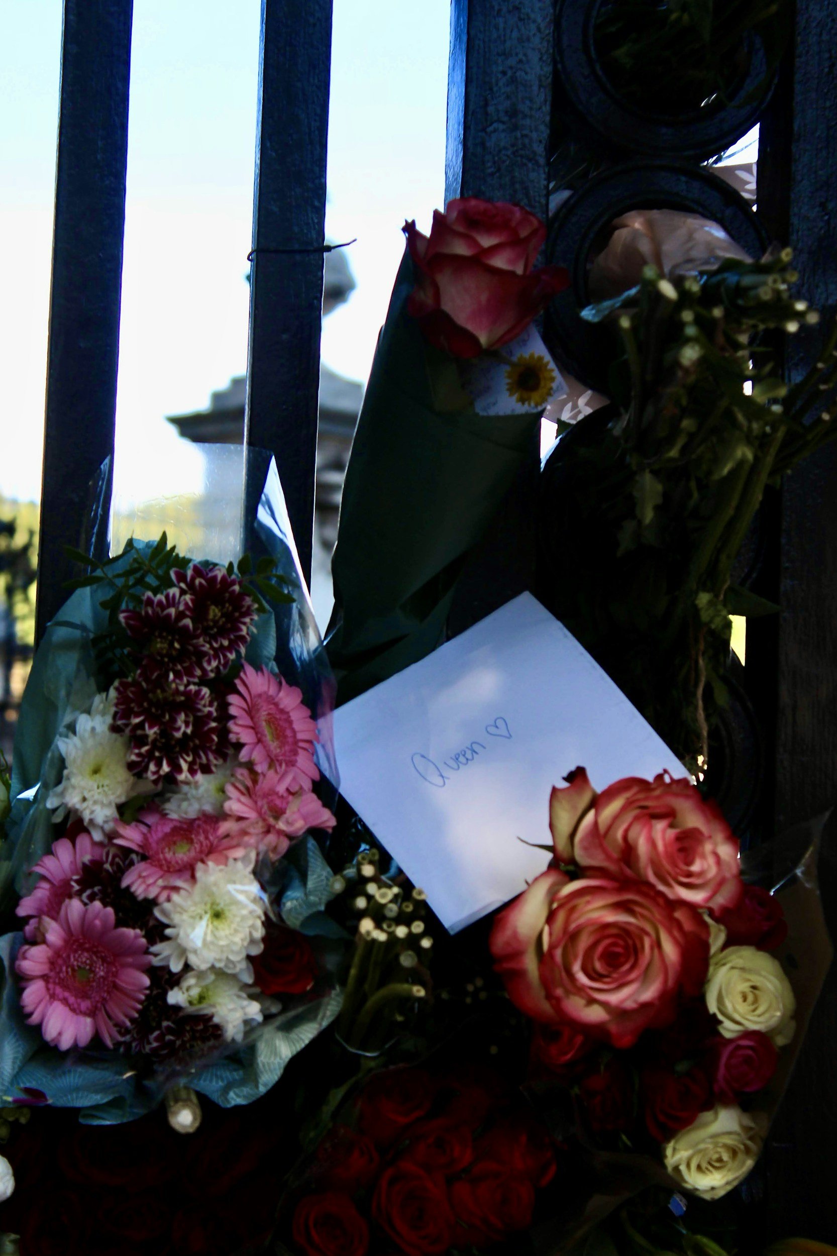Flowers and a handwritten note with the name 'Queen' and a heart on it, placed on a black fence.