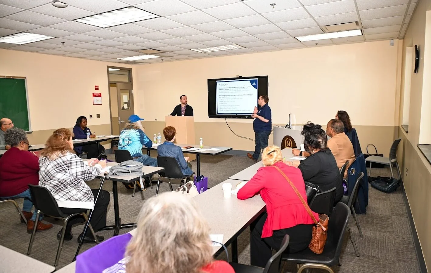 A classroom with people participating in a presentation. A man in a blue shirt is speaking in front of a large screen displaying a presentation slide. Attendees sit at desks, some taking notes, with papers, water bottles, and bags on the tables.