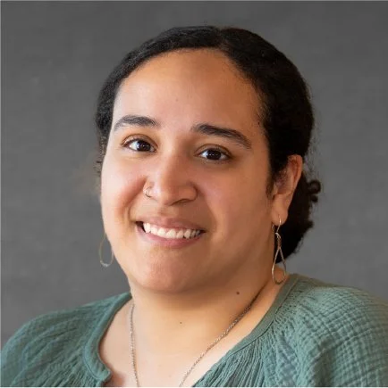 A smiling woman with dark hair tied back, wearing a green top, earrings, and a necklace, against a gray background.