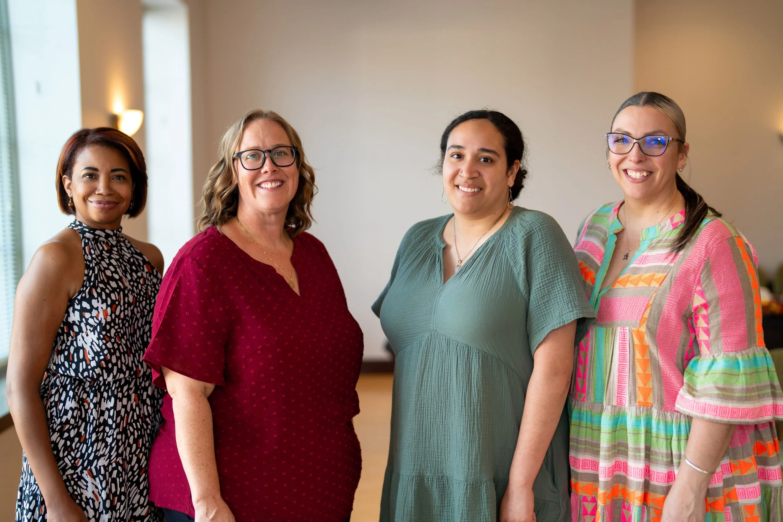 Four women standing together indoors, smiling at the camera, with a plain wall and windows in the background.