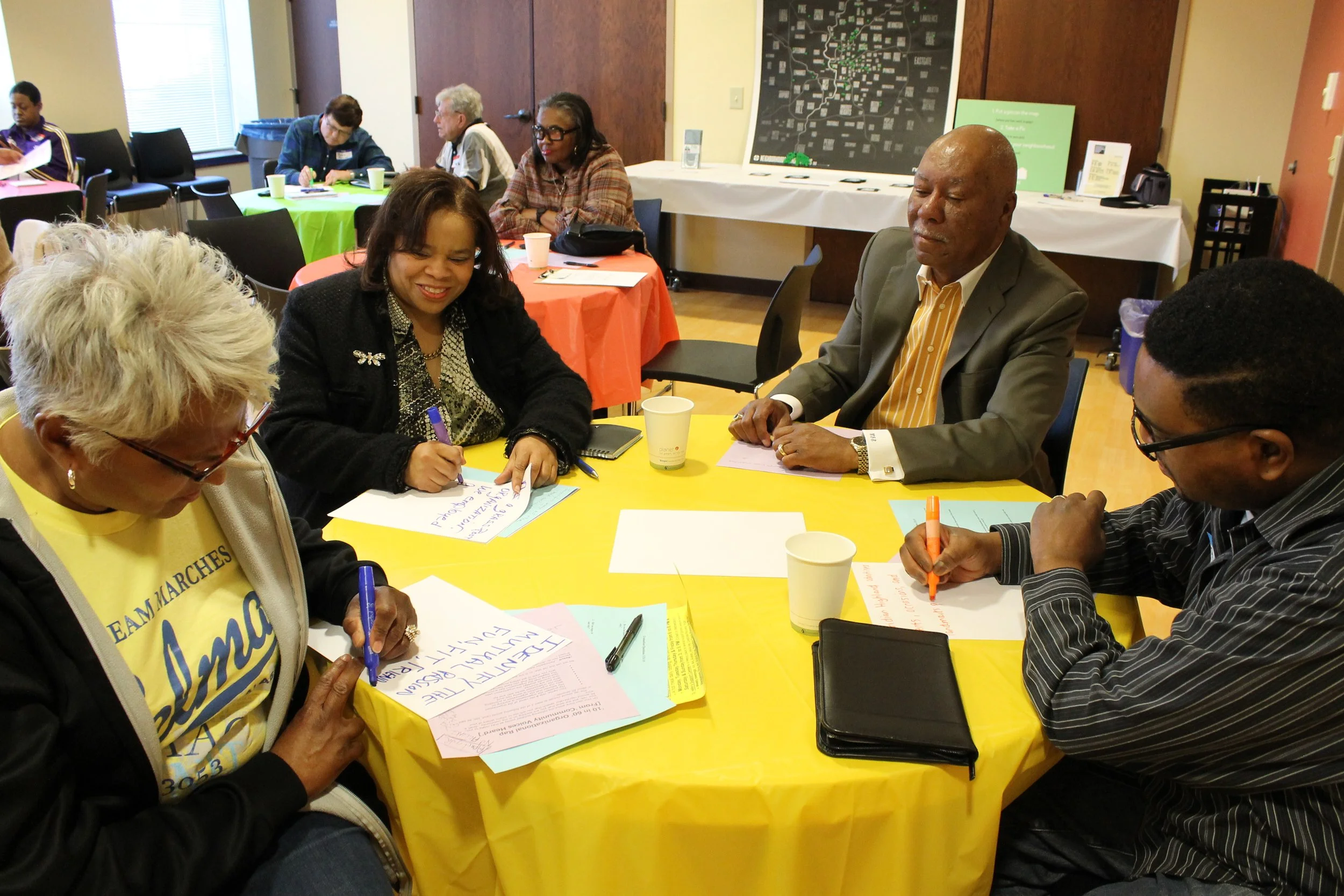 Group of five people sitting around a yellow table, engaged in a discussion during a workshop or meeting, with colorful tables and additional participants in the background.