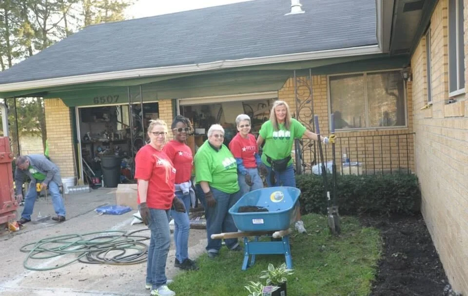 Group of volunteers planting flowers in a garden bed outside a yellow brick house.