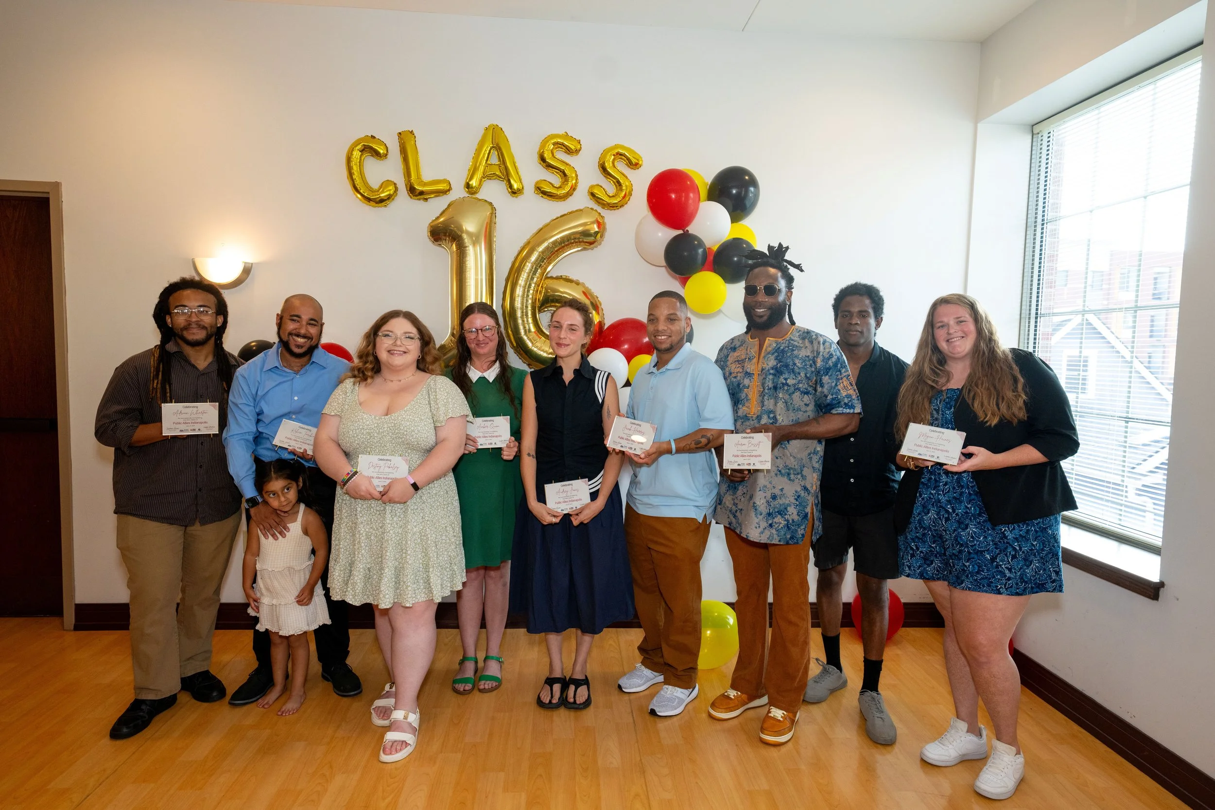 A group of ten people, including children, standing together in front of gold and black balloons, with large gold balloons spelling "CLASS" and a large gold "16" on the wall. They are celebrating a graduation or similar milestone, holding certificates, inside a room with a large window and hardwood floor.