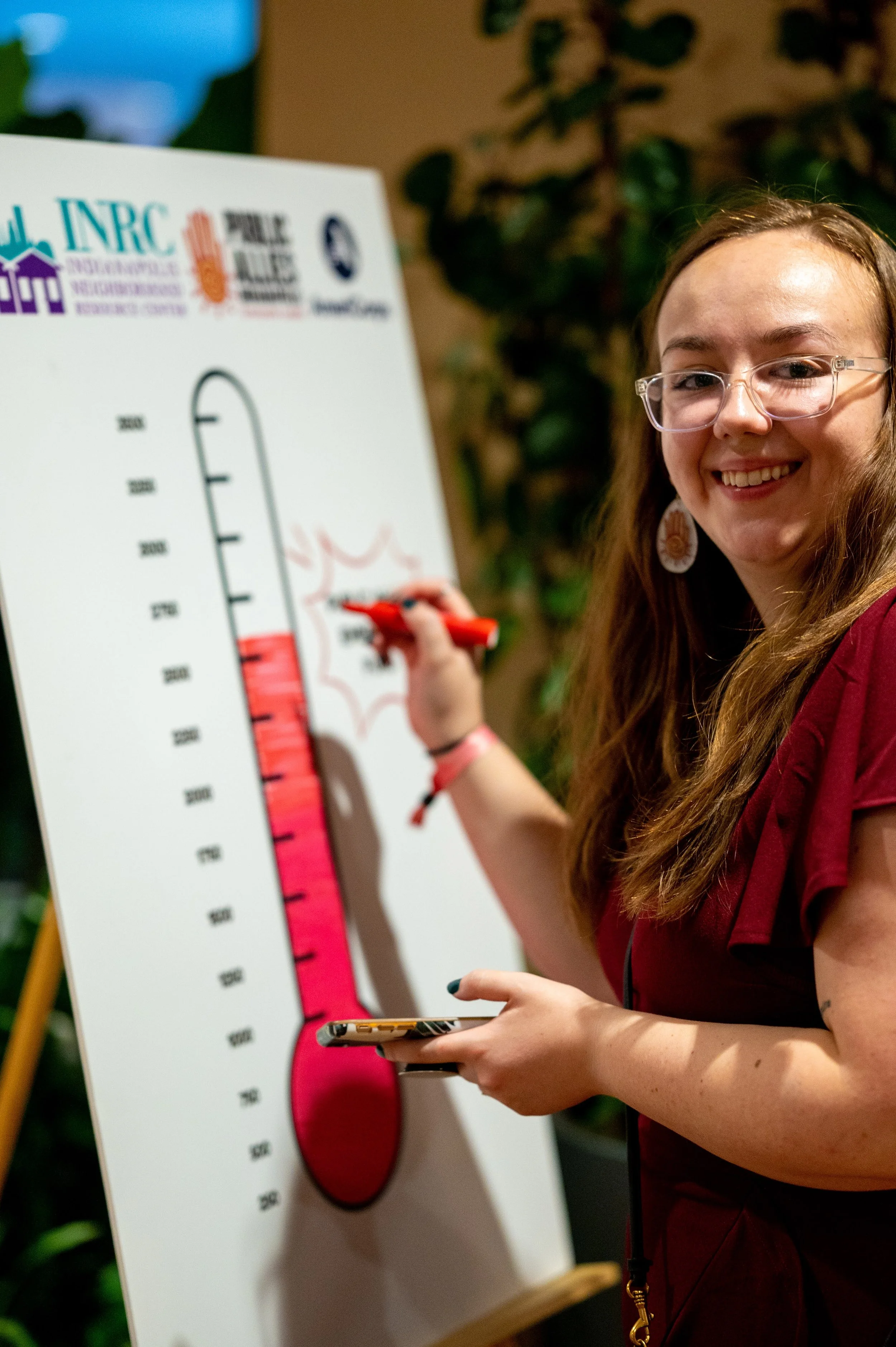 A smiling woman with glasses and long hair, holding a red marker, appears to be presenting a large thermometer chart on a whiteboard marking progress towards a fundraising goal.