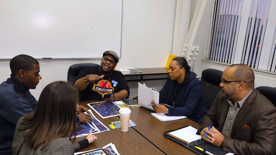Five people sitting around a conference table having a discussion, with some documents and a disposable coffee cup on the table, in an office setting.