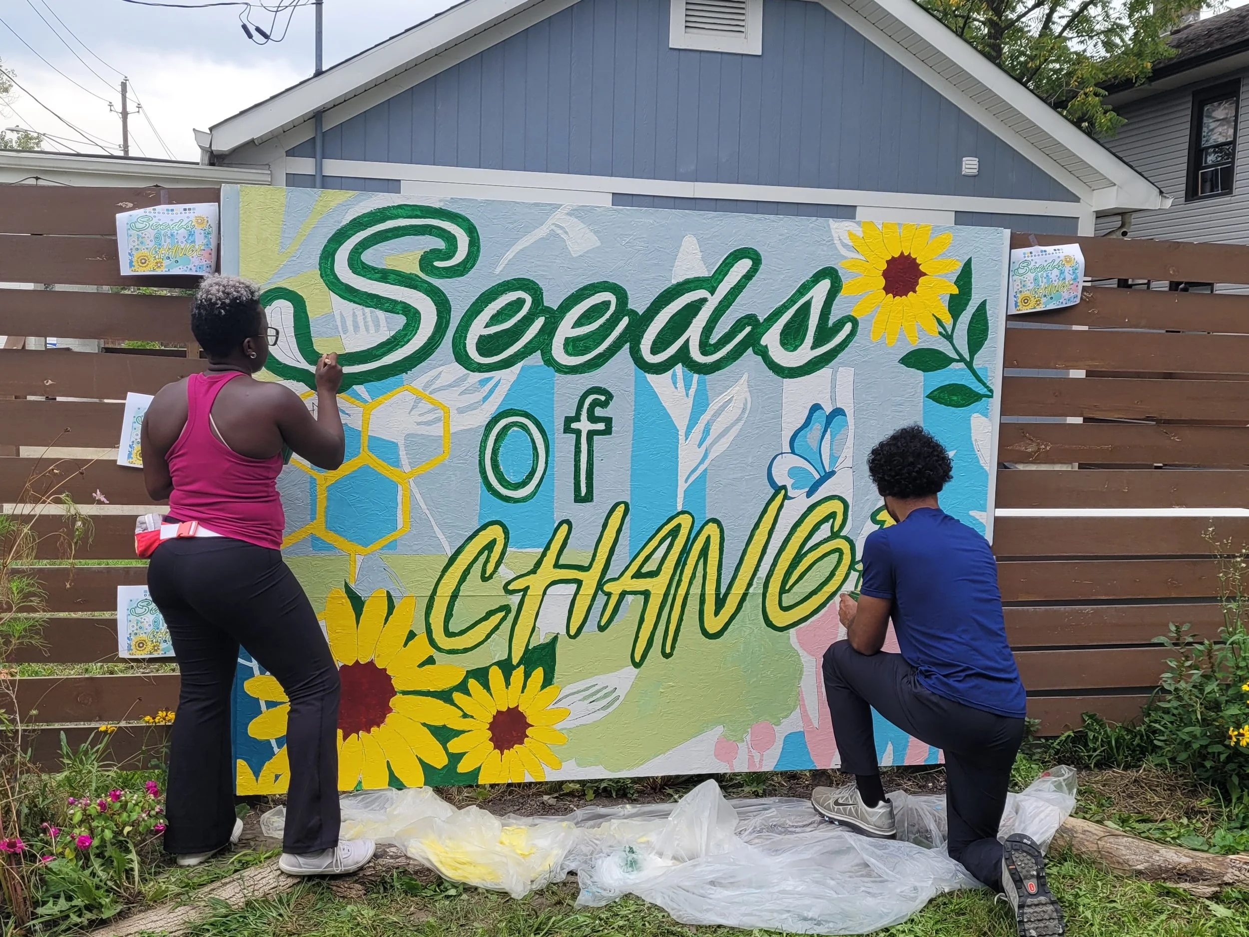 Two artists paint a colorful mural that says 'Seeds of Change' on a large outdoor wooden fence, with house and tree in background and art supplies on the ground.