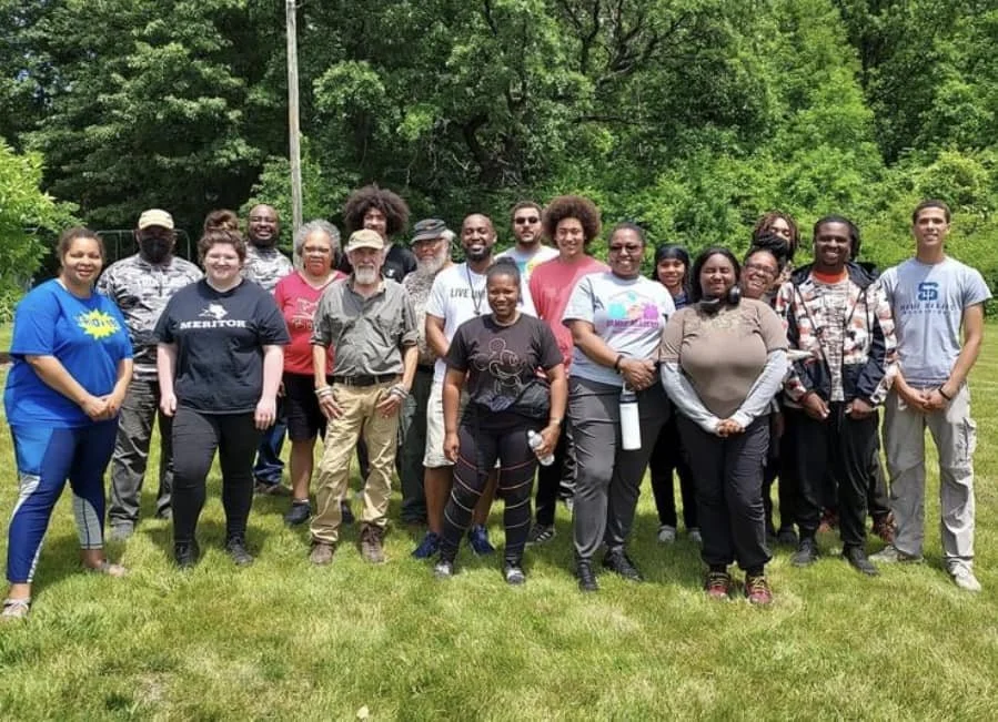 Group of diverse people posing outdoors on a grassy field with trees in the background.