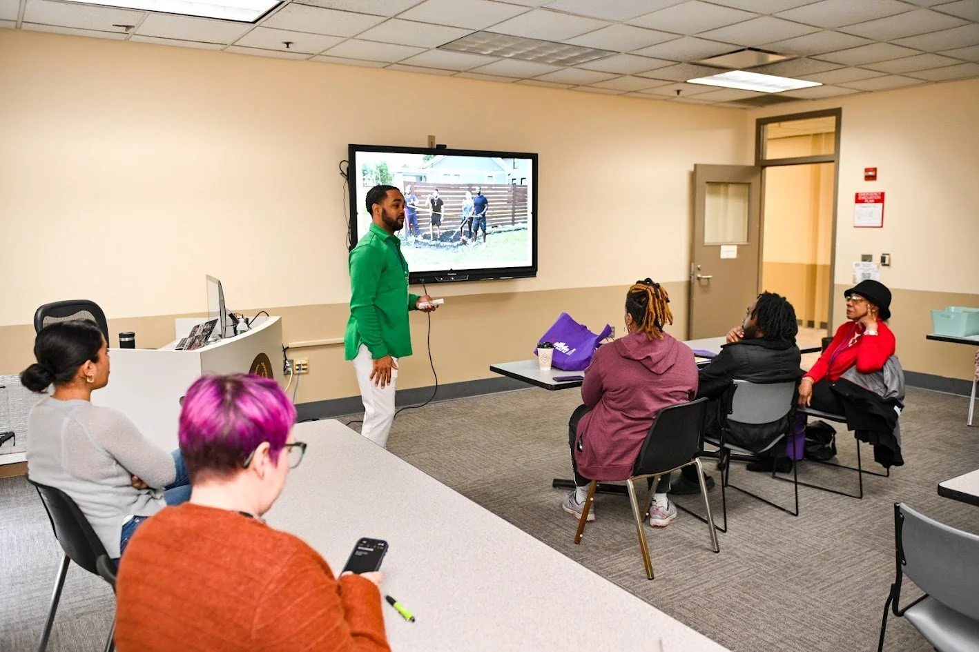 A man in a green shirt giving a presentation to a small group of women in a classroom or meeting room, with a large screen displaying an outdoor scene.