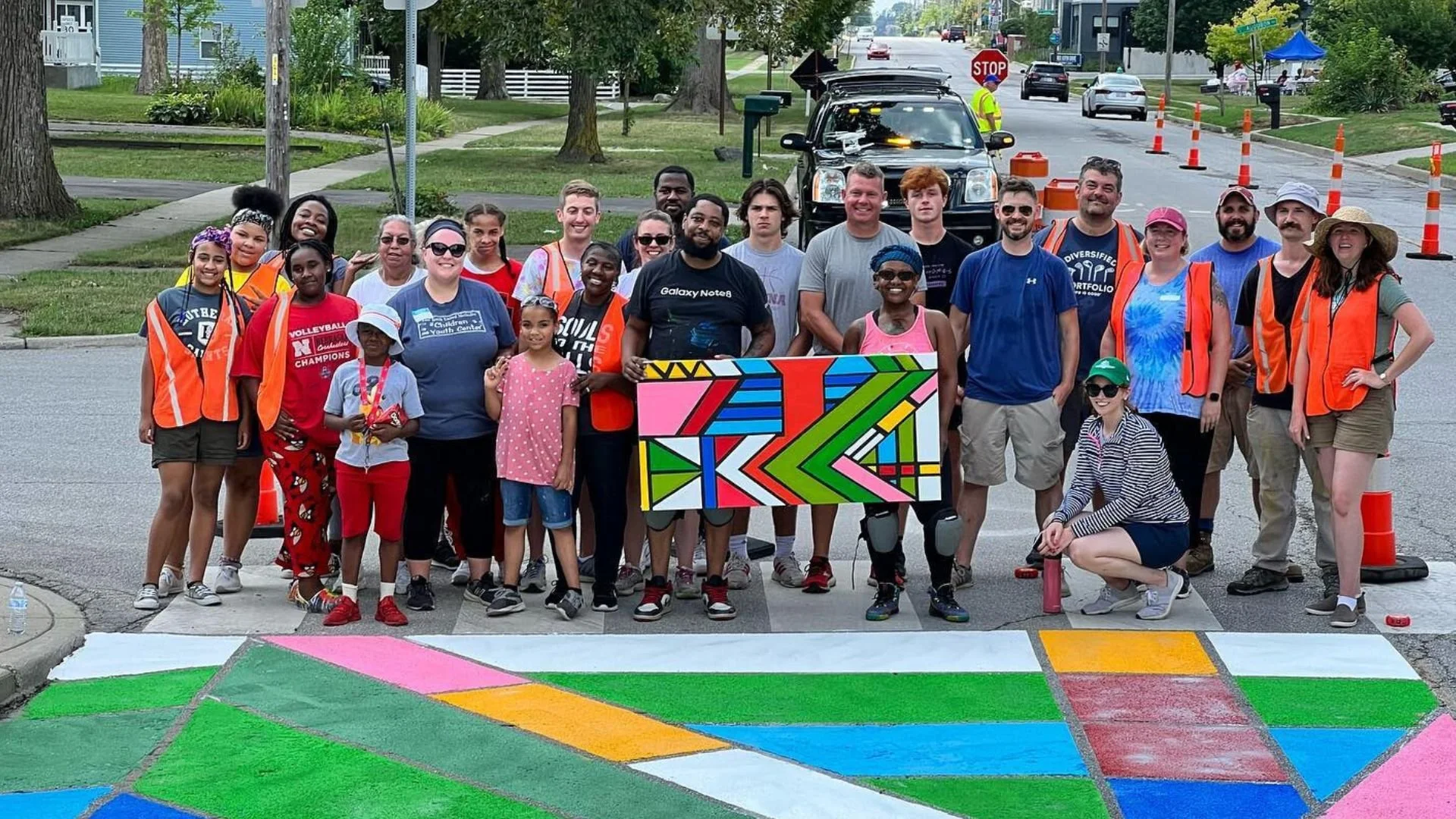 Group of diverse people, including children and adults, standing on a street corner with colorful street art. Some are wearing safety vests, and they are smiling for a group photo. Traffic cones and a black vehicle are visible in the background.