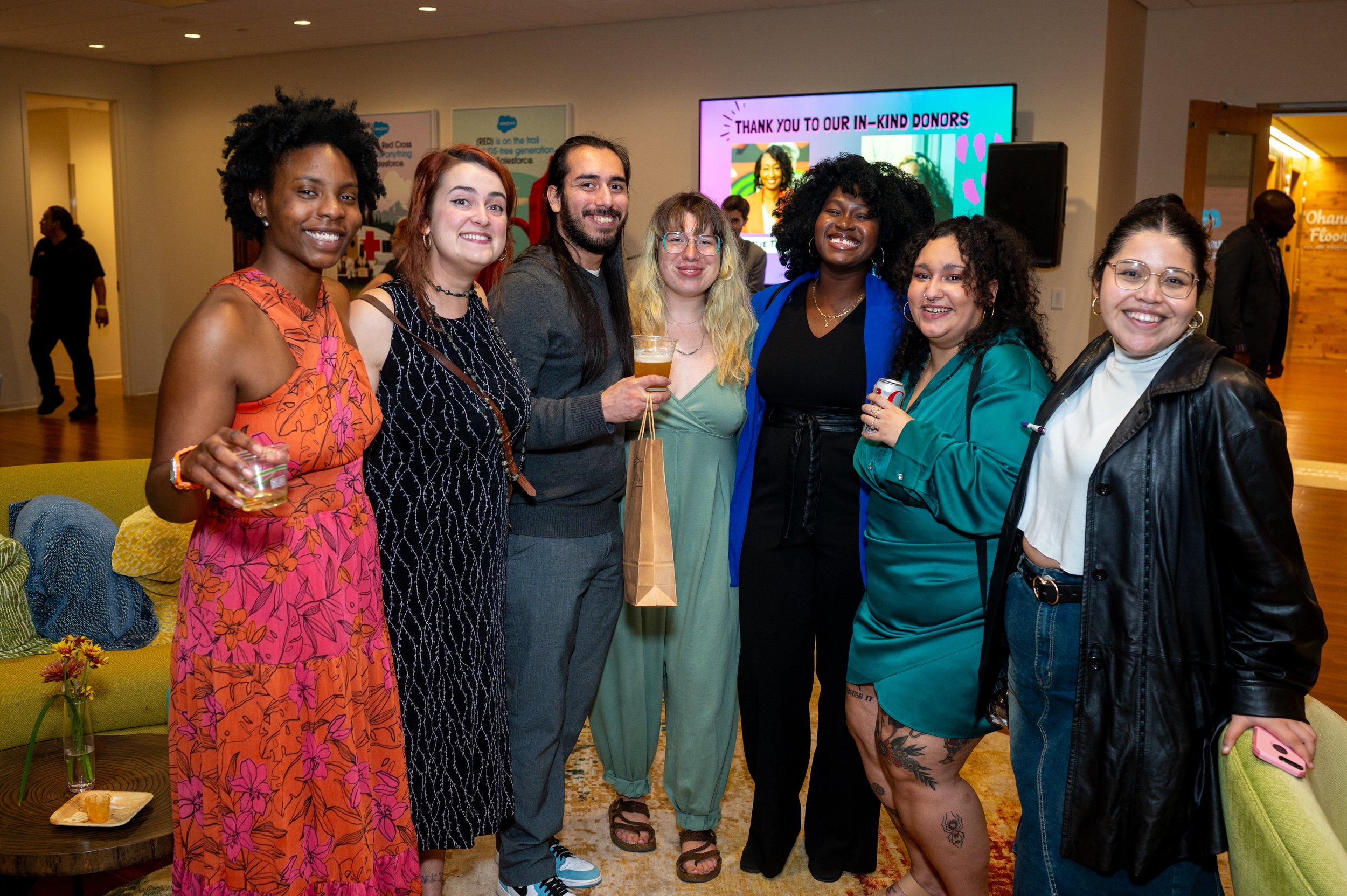 Group of seven diverse people smiling and posing together at an indoor event, holding drinks, with a colorful thank you message on a screen in the background.