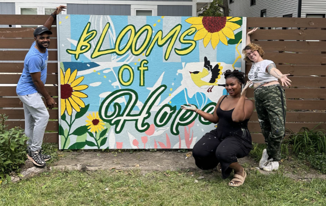 Three people pose in front of a colorful sign that reads 'Blooms of Hope' with sunflower illustrations, in an outdoor setting. One person stands on the left, another squats in the middle, and the third person stands on the right, all smiling and enjoying the moment.