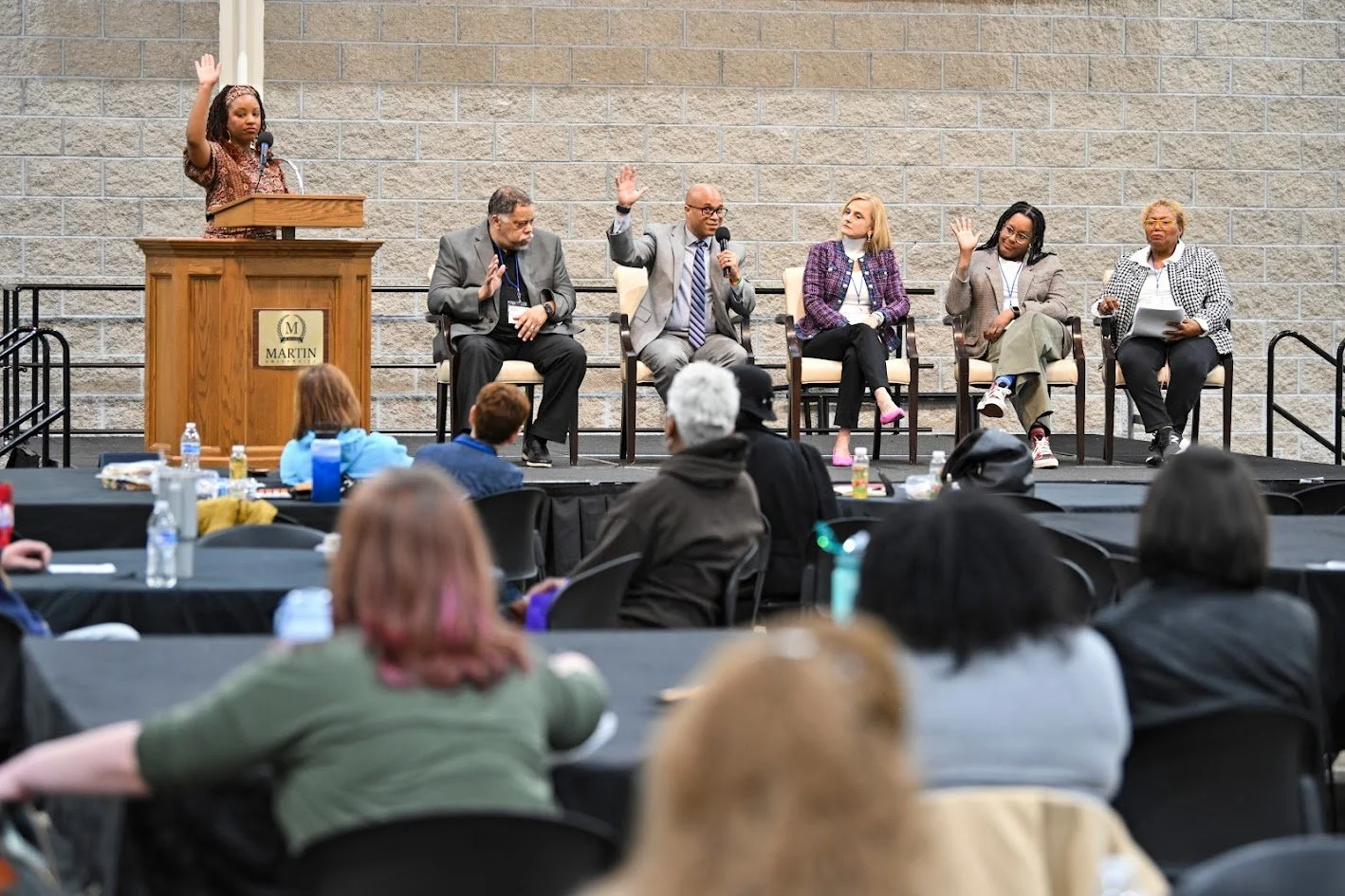A woman standing at a podium raising her hand during a panel discussion. Six adults seated behind her on stage, some also raising their hands. Audience members seated at tables in front.
