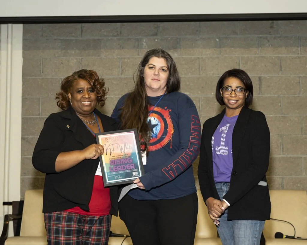 Three women standing indoors in front of a brick wall, one holding a framed certificate or award with the words 'Rising Leader' visible on it.