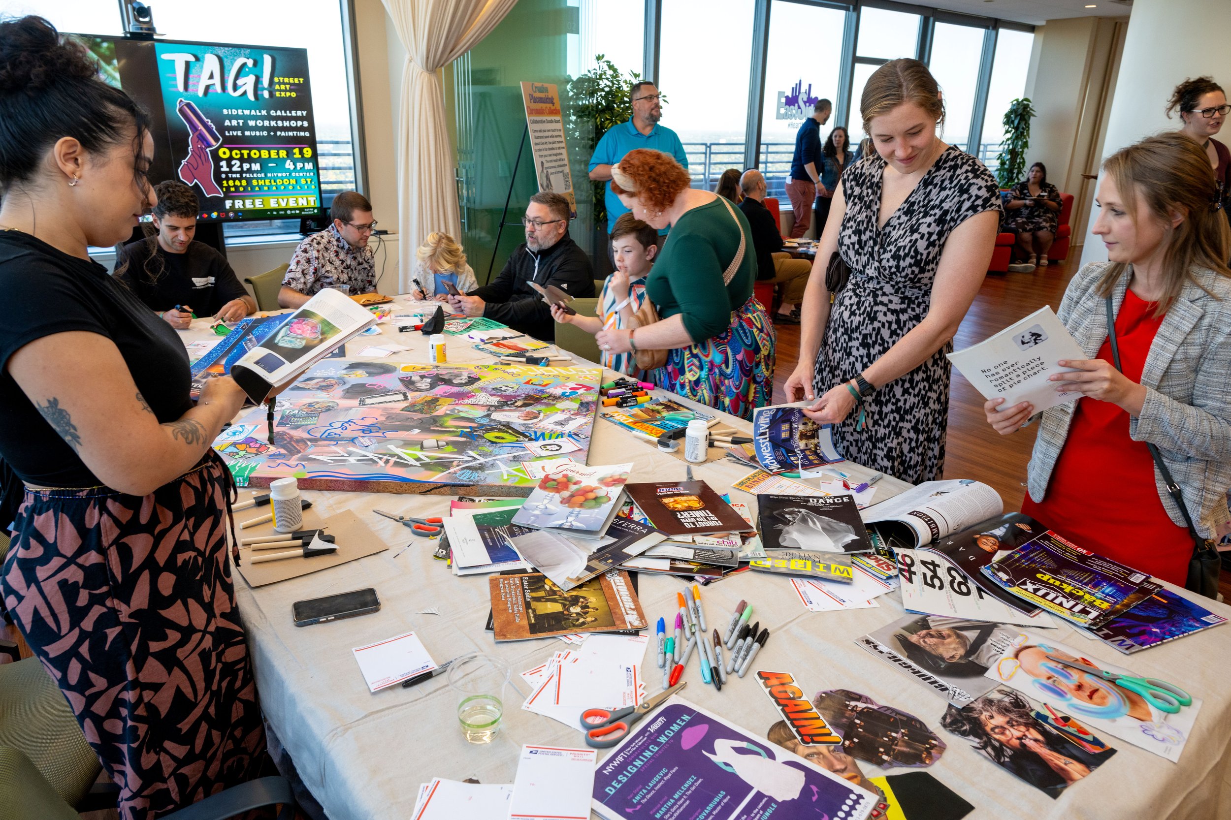 People gathered around a table working on creative projects, with art supplies, magazines, and scissors scattered on the table. In the background, a large digital screen displays an event poster.