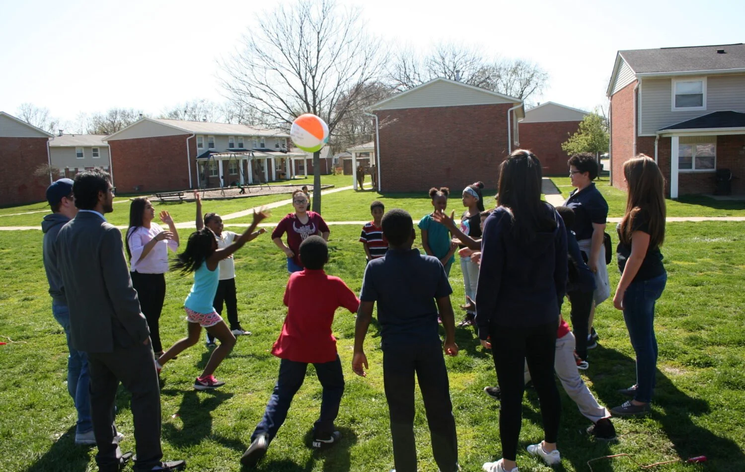 Group of children and adults playing with a beach ball outdoors on a grassy area in a residential neighborhood.