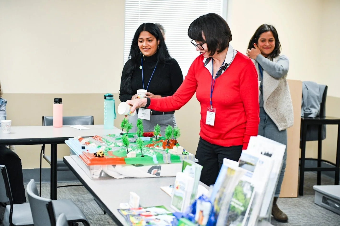 Three women at a conference or meeting, with a table displaying a miniature model landscape with trees, roads, and small vehicles.