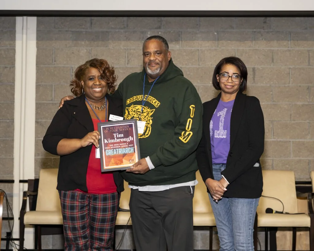 Three people standing together indoors, one woman holding a plaque, a man in the middle, and a woman on the right, all smiling. The woman on the left is wearing a black blazer and red top, the man is wearing a green hoodie with yellow lettering, and the woman on the right is dressed in a black blazer and purple shirt.
