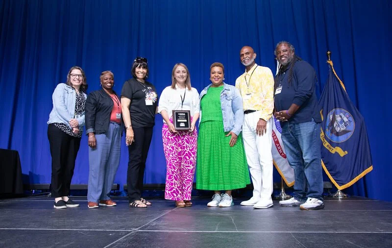 Seven people standing on stage in front of a blue curtain, some holding an award plaque. There are flags to the right, including one with an emblem. Everyone is smiling and dressed casually to semi-formally.