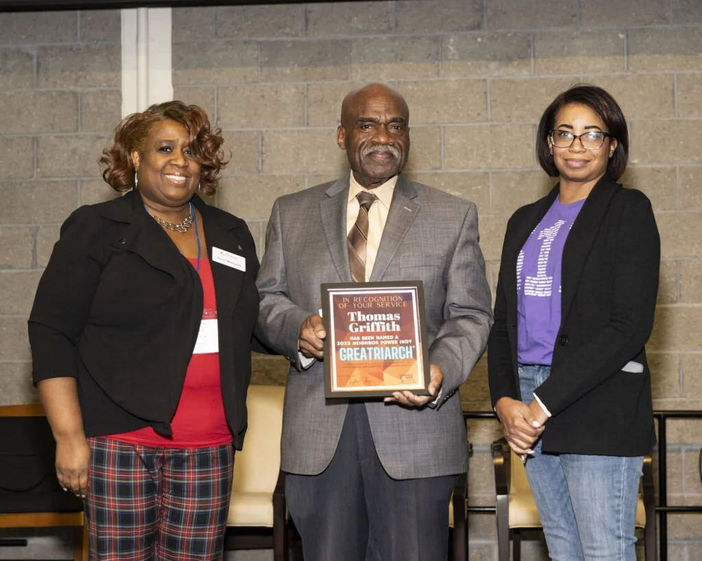 Three women and one man standing together indoors. The man in the center is holding a framed award plaque with the name Thomas Griffith and the title Greatriarch. The women on either side are smiling, with the woman on the left wearing a black jacket and plaid pants, and the woman on the right wearing glasses, a purple shirt, and a black blazer.