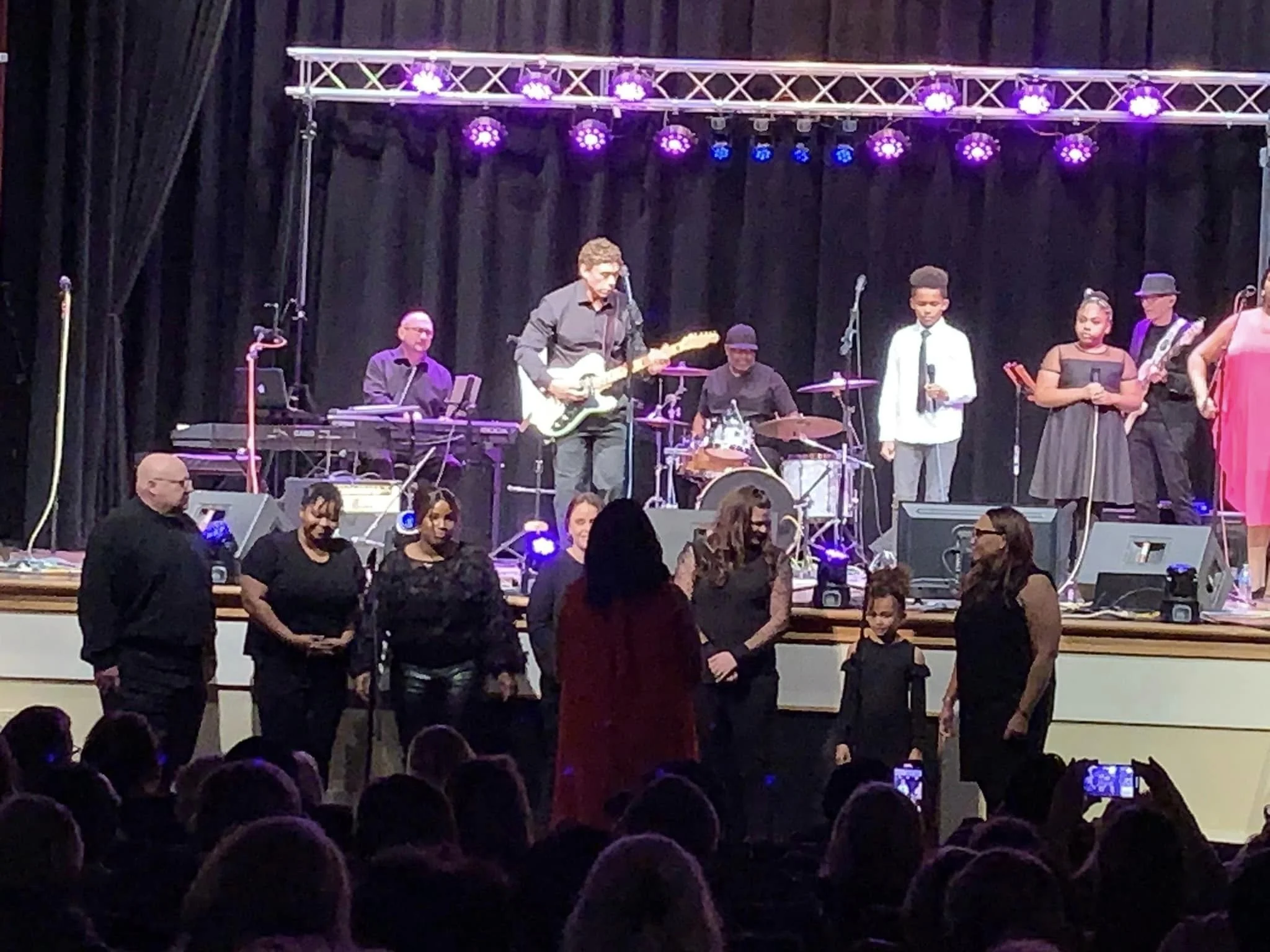 Musicians and singers performing on stage at a concert with purple lighting, with an audience in front taking pictures.