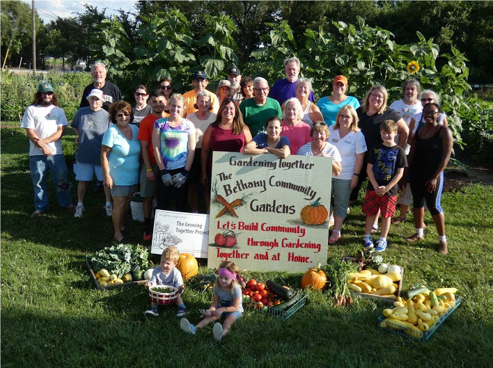 Group of people gathered in a garden holding a large sign that reads, "Gardening together, The Bethany Community Gardens. Let's Build Community through Gardening, Together and at Home." Kids and adults are present, with harvest baskets of vegetables and pumpkins in front, and large green plants in the background.