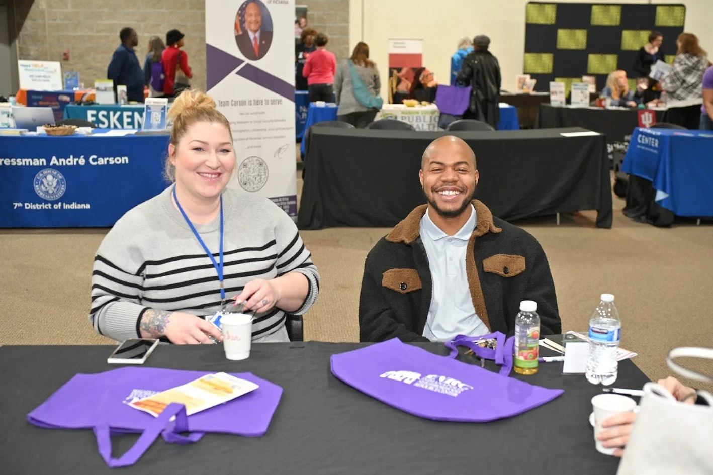 Two smiling people sitting at a table with purple bags on it at an indoor event. The woman on the left has blonde hair in a bun, wearing a gray and black striped sweater and a blue lanyard. The man on the right has a shaved head, wearing a black jacket with brown collar and a white shirt. There are water bottles, paper cups, and a smartphone on the table. Background shows booths and banners, including one for Congressman André Carson and the 7th District of Indiana.