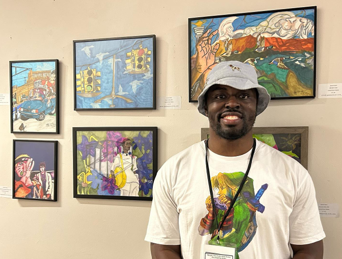 A smiling man wearing a gray bucket hat and a white t-shirt standing in front of a wall with colorful paintings displayed in an art gallery.