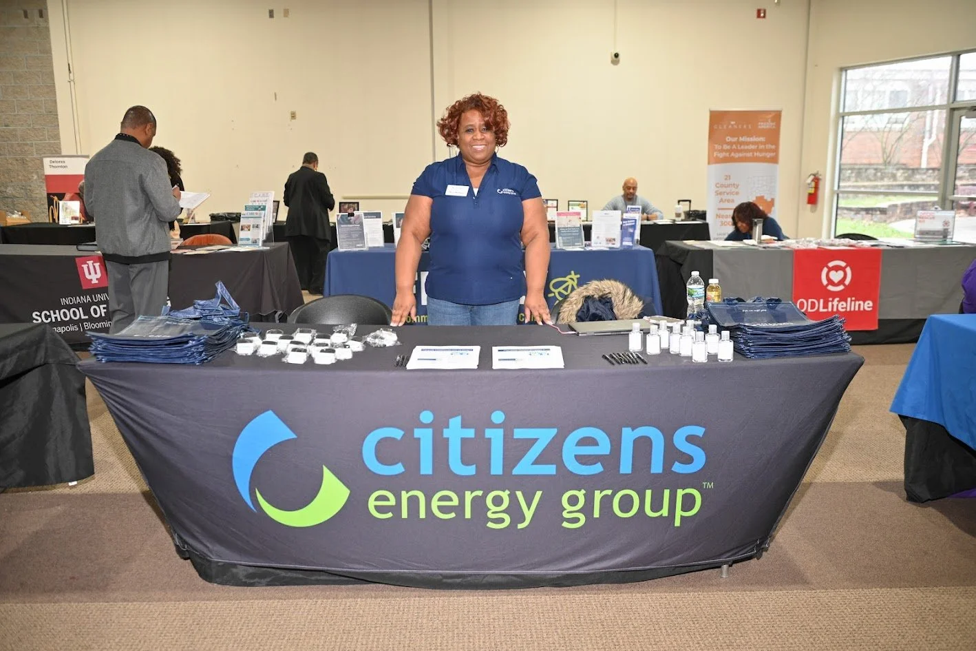 A woman standing behind a Citizens Energy Group booth at an indoor event, with various informational materials and small bottles on the table, in front of other booths and attendees.