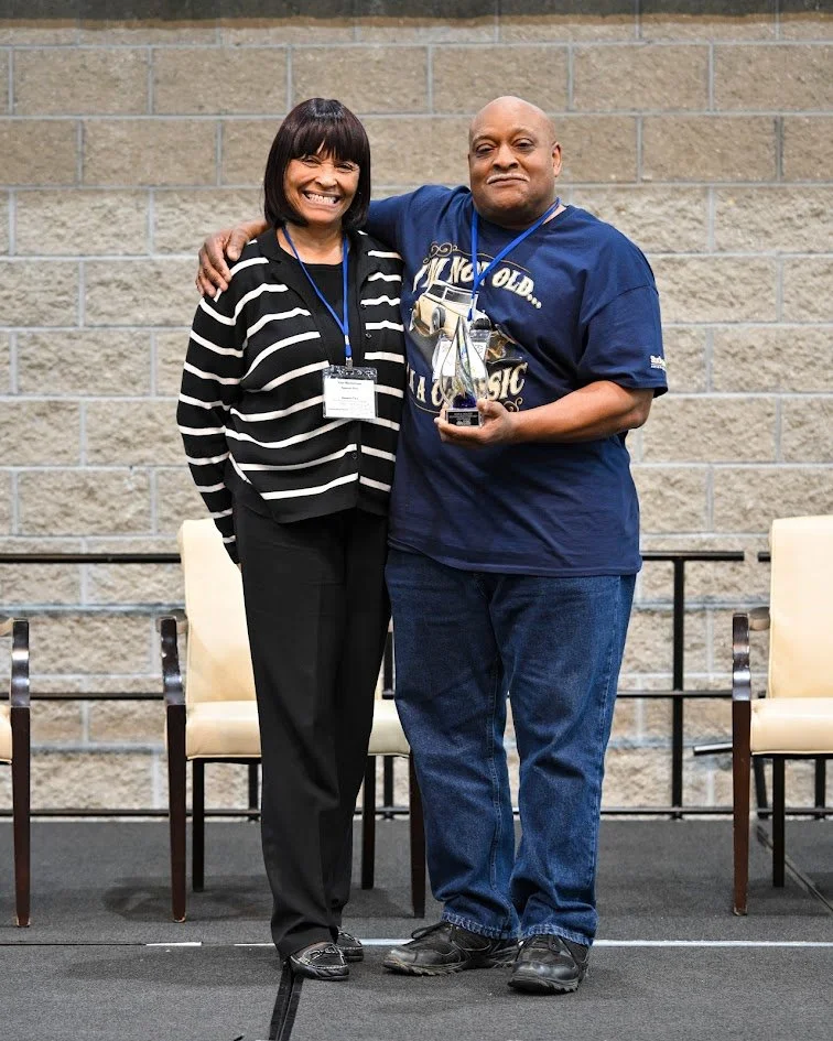 A woman and man standing together, smiling. The woman has shoulder-length dark hair and is wearing a black and white striped blazer, black pants, and black shoes. The man is bald, wearing a navy blue T-shirt with a graphic, dark jeans, and black shoes, holding a trophy. They are in front of a brick wall with empty chairs behind them.