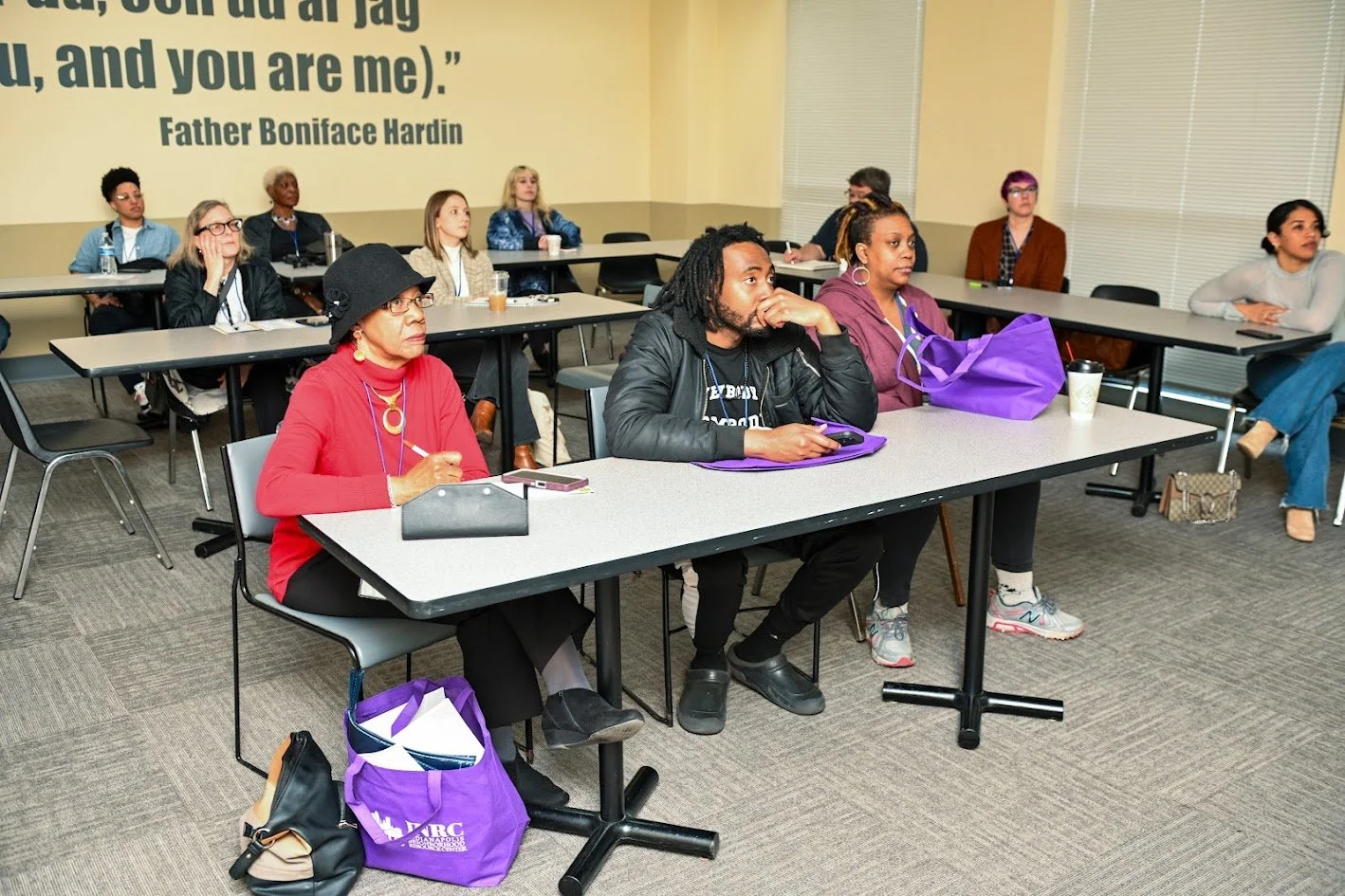 A group of people sitting at tables in a conference room, listening attentively. There are men and women of diverse ages and backgrounds, some with notebooks and purple tote bags on the table. A quote from Father Boniface Hardin is projected on the wall behind them.