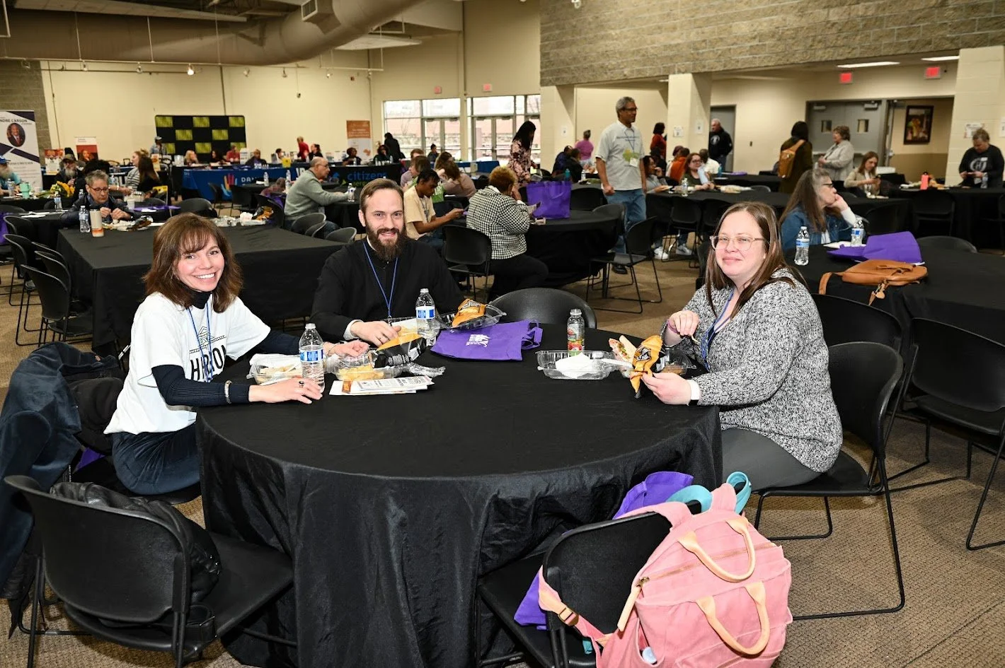 A group of three people sitting at a round table in a large conference room, eating lunch and smiling at the camera.