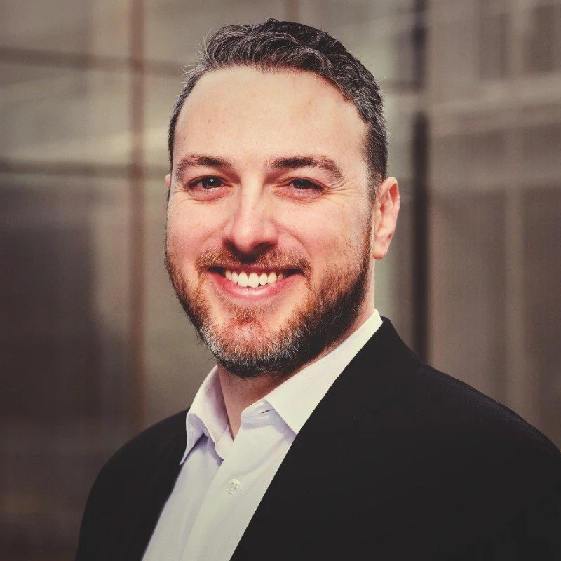 Portrait of a smiling man with a beard and short dark hair, wearing a black blazer and white shirt, in front of a blurred background.