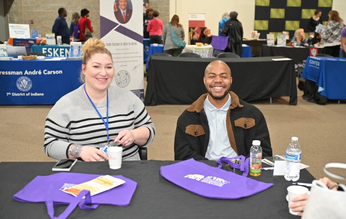 A woman and a man sitting at a table, smiling, with conference materials and water bottles in front of them, at an indoor event or conference.