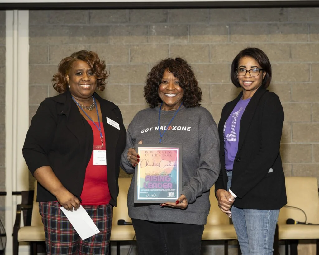 Three women smiling, standing indoors in front of a brick wall. The woman in the center is holding a framed award or certificate that says 'Rising Leader.'