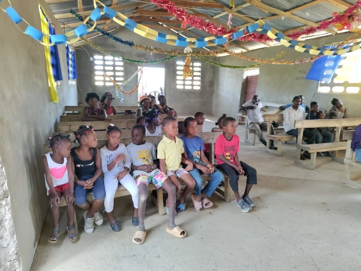 Children seated on wooden benches during a gathering in a classroom decorated with colorful paper garlands.