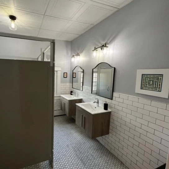 Two bathroom sinks with mirrors and light fixtures on light blue wall, white tiled lower wall, hexagonal tiled floor, and a privacy partition.