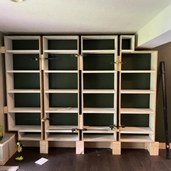 Wooden bookshelf framework in progress against a wall, with some outlets and construction tools nearby.