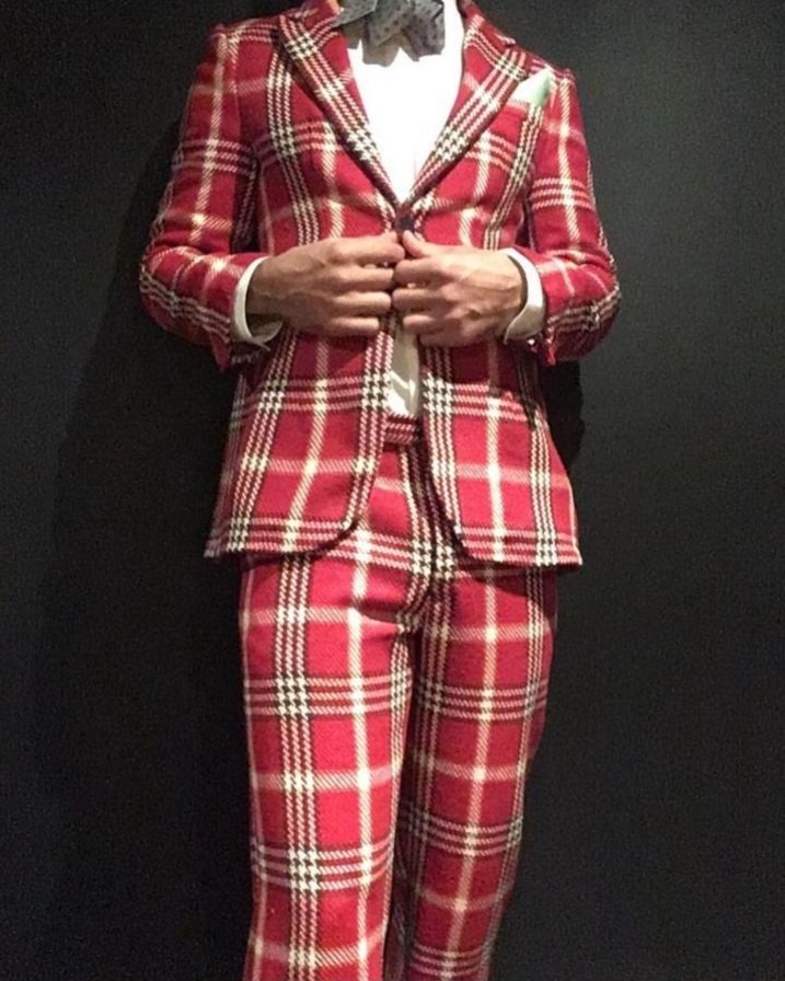 Person wearing a red tartan plaid suit with a matching blazer and pants, white shirt, and bow tie, standing against a black background.