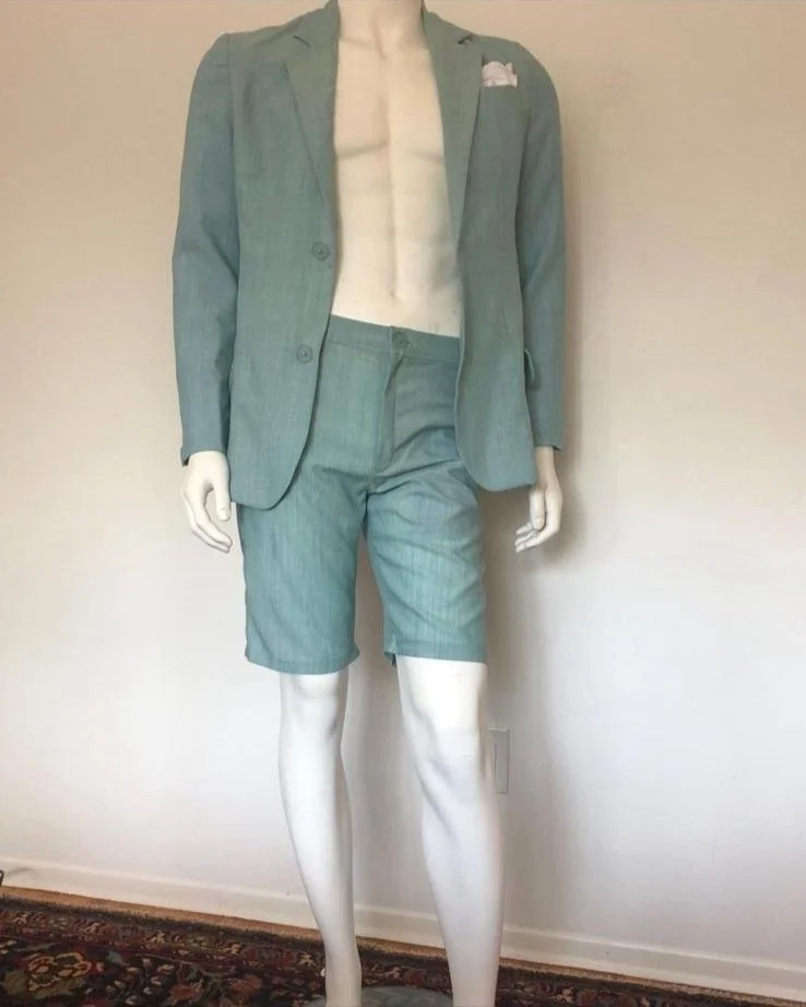 Mannequin dressed in a green business suit with blazer and shorts, exposed chest and arms, standing against a plain wall.