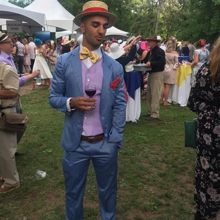 A man in a light blue suit, purple shirt, yellow bow tie, and straw hat holding a glass of red wine at an outdoor event with many people and tents in the background.