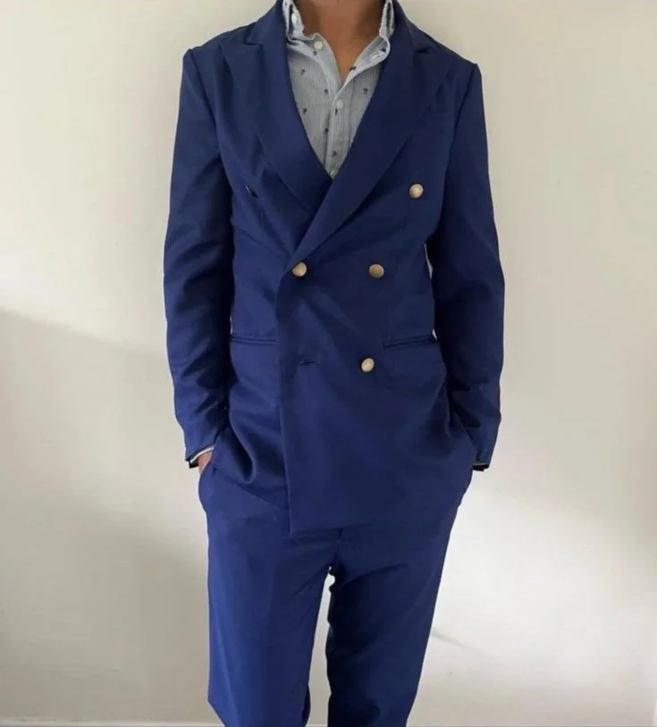 Man wearing a navy blue double-breasted suit with beige buttons, light blue dress shirt, and standing against a plain white wall.