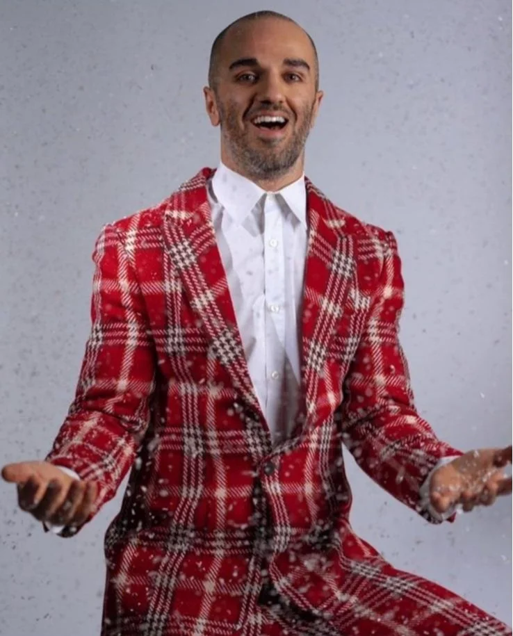 A man with a beard in a red plaid suit, smiling and gesturing with his hands, against a gray background with falling snow.