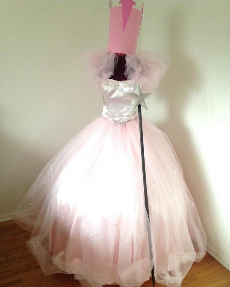 A mannequin dressed in a pink ball gown with a satin bodice, tulle sleeves, and a full layered tulle skirt, adorned with a star decoration, standing on a wooden floor against a plain wall.